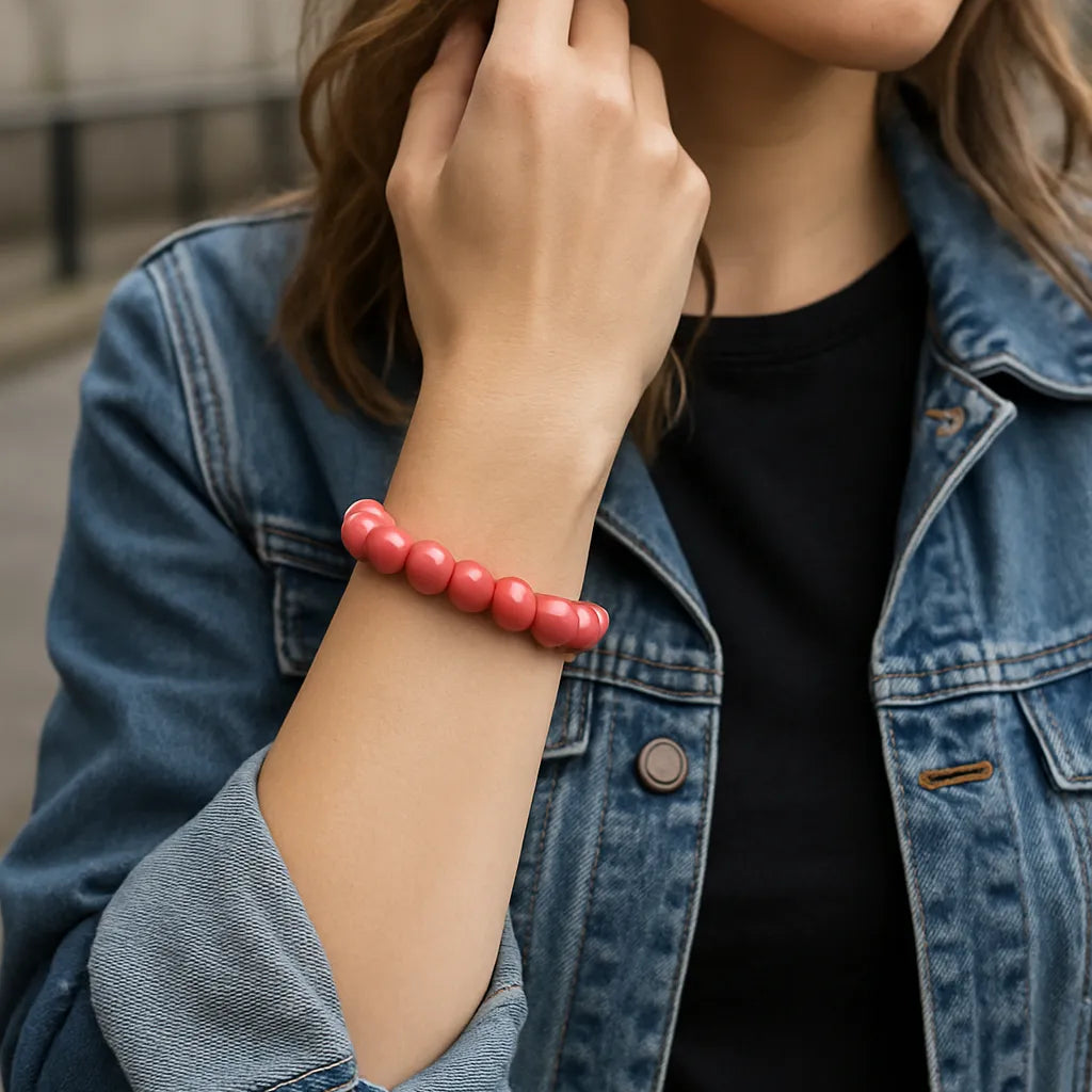 Eine junge Frau trägt ein lachsfarbenes Perlenarmband am Handgelenk. Sie steht in urbaner Umgebung, trägt eine Jeansjacke über einem schwarzen Shirt. Der Fokus liegt auf dem Armband.