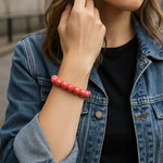 Eine junge Frau trägt ein lachsfarbenes Perlenarmband am Handgelenk. Sie steht in urbaner Umgebung, trägt eine Jeansjacke über einem schwarzen Shirt. Der Fokus liegt auf dem Armband.