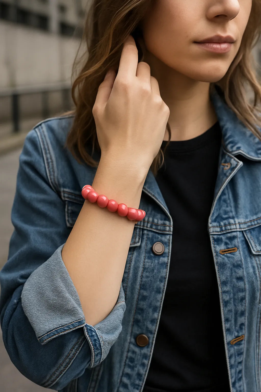 Eine junge Frau trägt ein lachsfarbenes Perlenarmband am Handgelenk. Sie steht in urbaner Umgebung, trägt eine Jeansjacke über einem schwarzen Shirt. Der Fokus liegt auf dem Armband.
