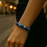 Eine Person trägt ein Armband aus glänzenden dunkelblauen und hellblauen Perlen am Handgelenk. Die Person steht auf einer Stadtstraße, trägt ein beigefarbenes Oberteil und schwarze Hose. Der Fokus liegt auf dem Armband und dem Unterarm.