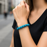 Eine Frau trägt ein türkisfarbenes Perlenarmband am Handgelenk. Der Fokus liegt auf dem Armband, während sie ihre Hand zum Gesicht hebt. Im Hintergrund ist eine unscharfe urbane Straße zu sehen.