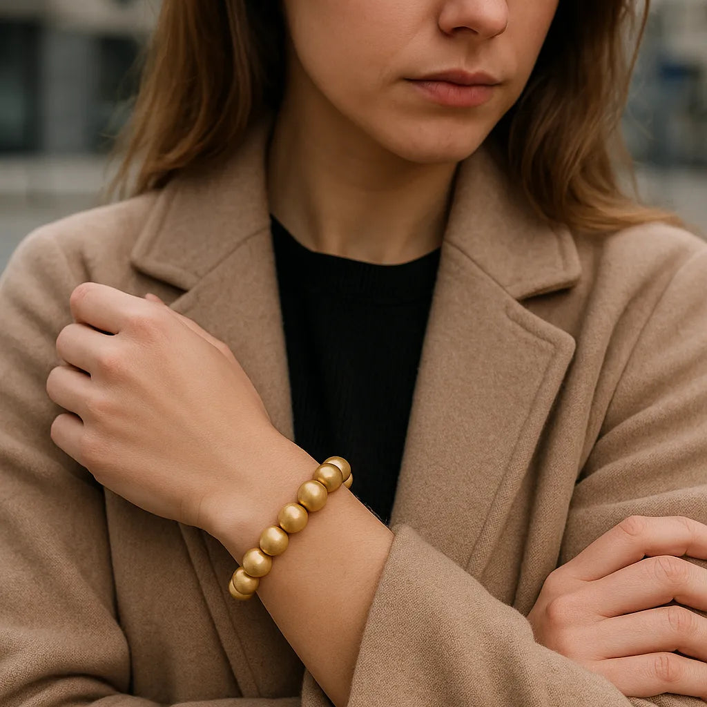 Eine Frau trägt ein goldenes Perlenarmband am Handgelenk. Sie steht in urbaner Umgebung, trägt einen beigefarbenen Mantel über einem schwarzen Oberteil. Der Fokus liegt auf dem Armband.