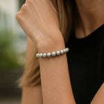 Eine Frau trägt ein silbernes Perlenarmband am Handgelenk. Der Fokus liegt auf dem Armband, während ihr Arm und ein schwarzes Oberteil im Vordergrund zu sehen sind. Der Hintergrund ist unscharf.