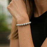 Eine Frau trägt ein silbernes Perlenarmband am Handgelenk. Der Fokus liegt auf dem Armband, während ihr Arm und ein schwarzes Oberteil im Vordergrund zu sehen sind. Der Hintergrund ist unscharf.