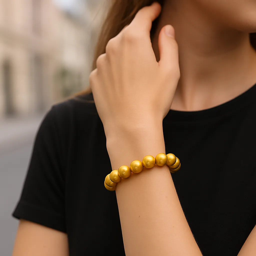 Eine Frau trägt ein goldenes Perlenarmband am Handgelenk. Der Fokus liegt auf dem Armband, während sie ihre Hand elegant zum Gesicht hebt. Im Hintergrund ist eine unscharfe urbane Umgebung erkennbar.