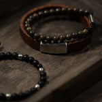 „Herren-Armbänder im Layering-Look: braunes geflochtenes Lederarmband mit Edelstahlverschluss und braunen Steinperlen plus schwarzes Perlenarmband auf Holz.“
